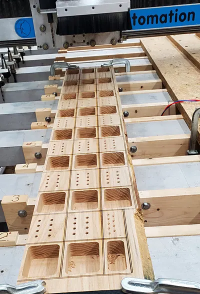 Hardwood board with a grid of CNC-cut pockets and drilled holes, clamped on the router